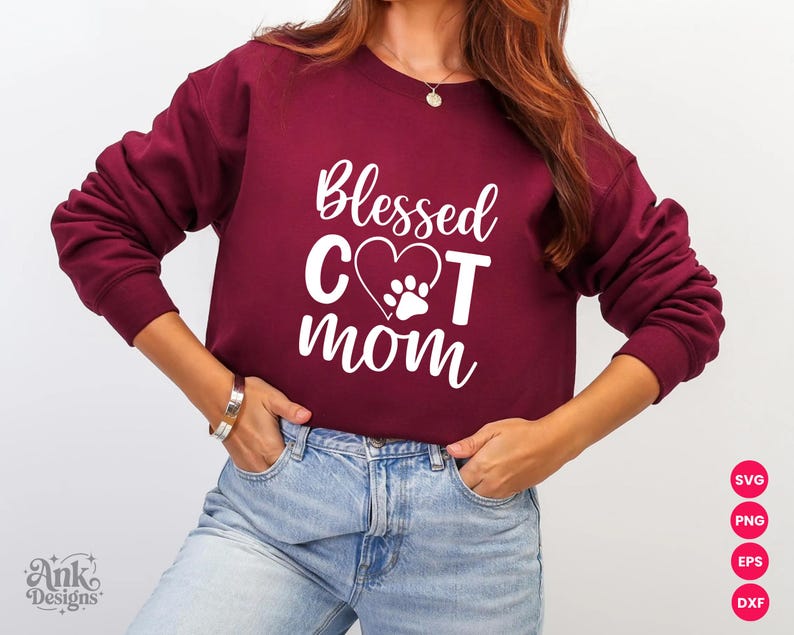 以下が含まれることがあります： 「Blessed Cat Mom」の白文字がプリントされた、マルーン色のスウェットシャツ。デザインには、足跡が入ったハートが含まれています。スウェットシャツはクルーネックです。