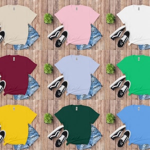 K&ouml;nnte beinhalten: Neun einfarbige T-Shirts in Beige, Rosa, Wei&szlig;, Burgunderrot, Hellblau, Gr&uuml;n, Gelb, Dunkelgr&uuml;n und Hellblau. Jedes Shirt wird mit Jeansshorts und schwarzen Turnschuhen auf einem Holzgrund gezeigt.