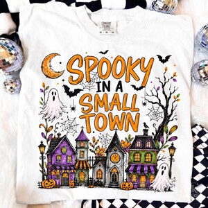 Può includere: Una t-shirt bianca con una stampa grafica che riporta il testo "SPOOKY IN A SMALL TOWN" in lettere arancioni. Il design include una luna crescente, pipistrelli, ragni, ragnatele, due fantasmi e una fila di case colorate.