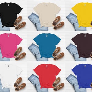 K&ouml;nnte beinhalten: Neun einfarbige T-Shirts in Schwarz, Beige, Gelb, Pink, T&uuml;rkis, Burgunderrot, Wei&szlig;, Rot und Blau. Jedes Shirt wird mit blauen Jeans und braunen Turnschuhen auf einer wei&szlig;en Holzoberfl&auml;che pr&auml;sentiert.