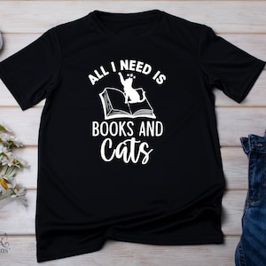 以下が含まれることがあります： 「ALL I NEED IS BOOKS AND CATS」と白い文字で書かれた黒いTシャツ。デザインは猫と開いた本です。Tシャツは明るい木製の表面に表示され、ジーンズと花が近くにあります。