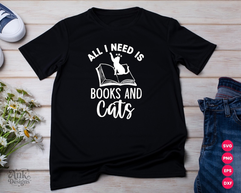 以下が含まれることがあります： 「ALL I NEED IS BOOKS AND CATS」と白い文字で書かれた黒いTシャツ。デザインは猫と開いた本です。Tシャツは明るい木製の表面に表示され、ジーンズと花が近くにあります。