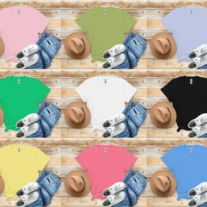 K&ouml;nnte beinhalten: Eine Sammlung von neun farbigen T-Shirts in Rosa, Gr&uuml;n, Blau, Wei&szlig;, Schwarz, Gelb und Koralle. Jedes Shirt ist mit Jeans, einem braunen Hut und wei&szlig;en Turnschuhen auf Holzgrund angeordnet.