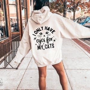 以下が含まれることがあります： 「I ONLY HAVE eyes for MY CATS」の黒い文字が入ったクリーム色のフード付きスウェットシャツ。猫の目と星のデザイン。スウェットシャツは屋外で着用されています。