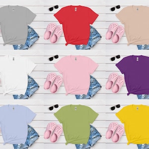 K&ouml;nnte beinhalten: Neun einfarbige T-Shirts in verschiedenen Farben: Grau, Rot, Beige, Wei&szlig;, Rosa, Lila, Hellblau, Gr&uuml;n und Gelb. Jedes Shirt wird mit Jeansshorts, rosa Clogs und Sonnenbrillen auf einer wei&szlig;en Holzoberfl&auml;che pr&auml;sentiert.