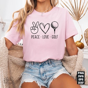 May include: A light pink t-shirt with the words "PEACE LOVE GOLF" in black. The design includes a peace sign, a heart, and a golf ball on a tee. The shirt is a casual style.