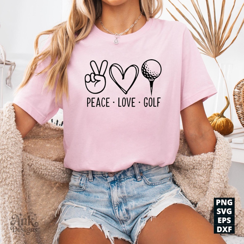 May include: A light pink t-shirt with the words "PEACE LOVE GOLF" in black. The design includes a peace sign, a heart, and a golf ball on a tee. The shirt is a casual style.