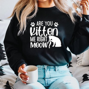 以下が含まれることがあります： 「ARE YOU kitten ME RIGHT MEOW？」の白文字が入った黒いスウェットシャツ。足跡と猫のシルエットのデザイン。スウェットシャツはブルージーンズと合わせています。白いマグカップが手に持たれています。