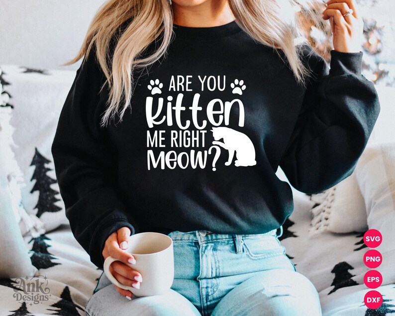 以下が含まれることがあります： 「ARE YOU kitten ME RIGHT MEOW？」の白文字が入った黒いスウェットシャツ。足跡と猫のシルエットのデザイン。スウェットシャツはブルージーンズと合わせています。白いマグカップが手に持たれています。