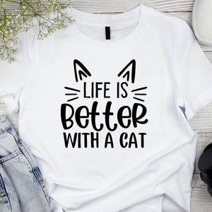 以下が含まれることがあります： 「LIFE IS BETTER WITH A CAT」の黒い文字がプリントされた白いTシャツ。猫の耳とひげが特徴です。明るい色の表面に、ジーンズ、白いスニーカー、ヘッドフォンと一緒に表示されています。