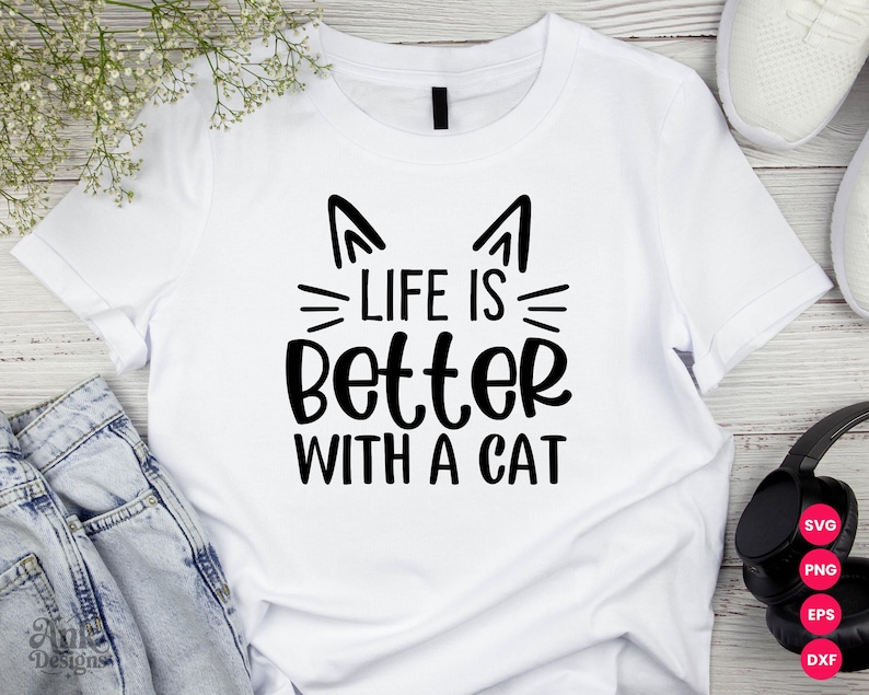 以下が含まれることがあります： 「LIFE IS BETTER WITH A CAT」の黒い文字がプリントされた白いTシャツ。猫の耳とひげが特徴です。明るい色の表面に、ジーンズ、白いスニーカー、ヘッドフォンと一緒に表示されています。