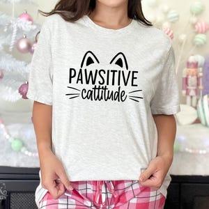 以下が含まれることがあります： 「PAWSITIVE cattitude」の黒い文字がプリントされたライトグレーのTシャツ。猫の耳とヒゲのグラフィックデザイン。ピンクと白のチェック柄のパジャマパンツと組み合わせています。猫をテーマにしたデザイン。