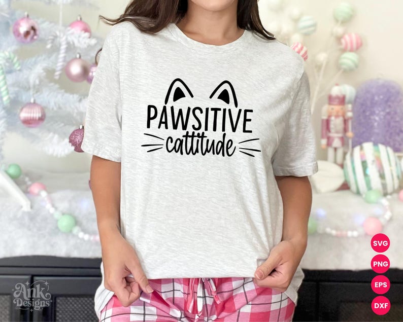 以下が含まれることがあります： 「PAWSITIVE cattitude」の黒い文字がプリントされたライトグレーのTシャツ。猫の耳とヒゲのグラフィックデザイン。ピンクと白のチェック柄のパジャマパンツと組み合わせています。猫をテーマにしたデザイン。