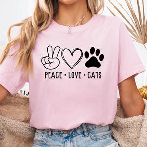 以下が含まれることがあります： 黒いグラフィックデザインの明るいピンク色のTシャツ。デザインには平和のサイン、ハート、猫の足跡が含まれており、下に「PEACE LOVE CATS」という言葉があります。シャツは柔らかい素材で作られています。
