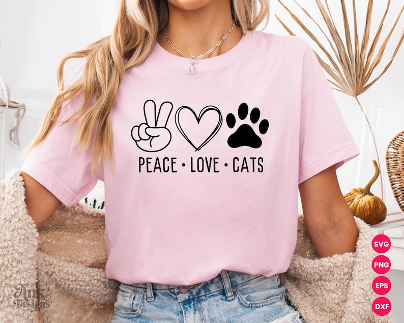 以下が含まれることがあります： 黒いグラフィックデザインの明るいピンク色のTシャツ。デザインには平和のサイン、ハート、猫の足跡が含まれており、下に「PEACE LOVE CATS」という言葉があります。シャツは柔らかい素材で作られています。