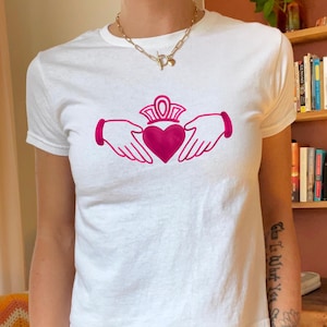 May include: White t-shirt with a pink Claddagh design. The Claddagh symbol, featuring a heart, crown, and hands, is printed on the front. The shirt has short sleeves and a round neck.