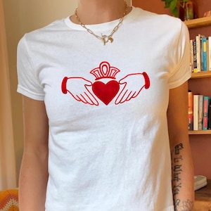 Puede incluir: Camiseta blanca con un diseño Claddagh rojo. El símbolo de Claddagh, con un corazón, una corona y manos, está centrado en la camiseta. La camiseta tiene mangas cortas y cuello redondo.