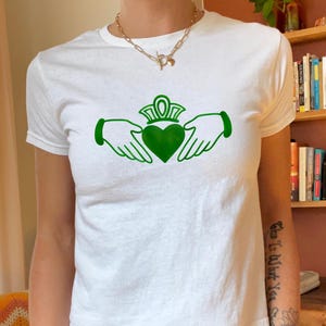 Puede incluir: Camiseta blanca con un diseño Claddagh verde. El símbolo de Claddagh, con un corazón, una corona y manos, está impreso en la parte delantera. La camiseta es de cuello redondo.