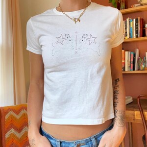 以下が含まれることがあります： クルーネックと半袖の白いTシャツ。Tシャツには、2匹のウサギと星の対称的なデザインが施されています。モデルはブルージーンズとゴールドのネックレスを着用しています。