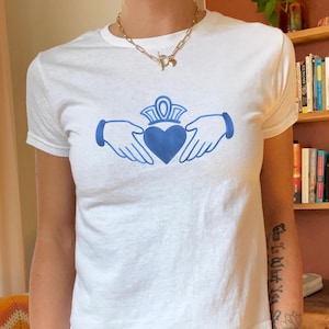 May include: White t-shirt featuring a blue Claddagh design. The Claddagh symbol, with a heart, crown, and hands, is centered on the shirt. The shirt has short sleeves and a crew neck.