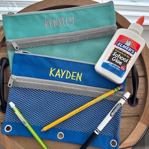 May include: A teal zippered pouch embroidered with the name KENSLEY and a blue zippered pouch embroidered with the name KAYDEN, both featuring mesh fronts. Included are a bottle of Elmer's School Glue, a yellow pencil, a green pencil, and a marker.
