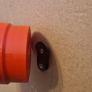 May include: A black plastic wall mount with two screws. The mount is attached to a beige wall. A red pipe is visible to the left of the mount.
