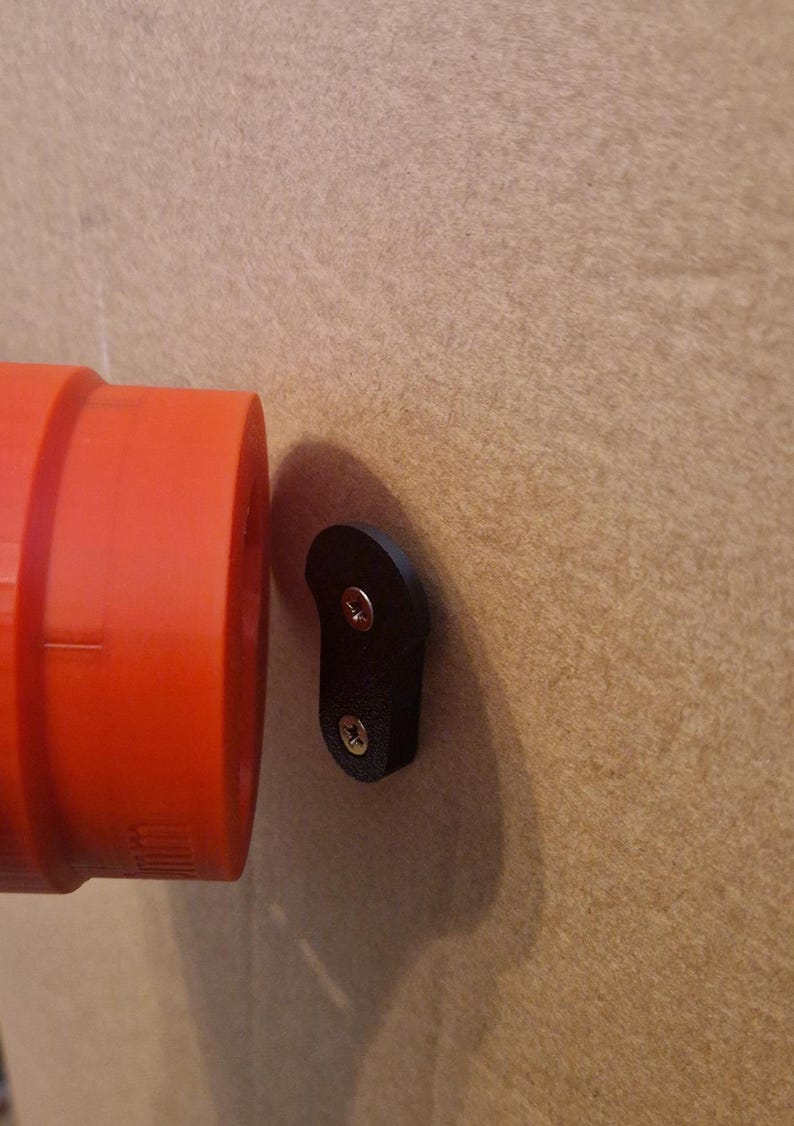 May include: A black plastic wall mount with two screws. The mount is attached to a beige wall. A red pipe is visible to the left of the mount.