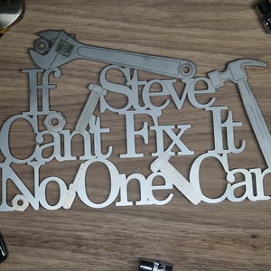 May include: A silver metal sign with the text "If Steve Can't Fix It No One Can" and tool silhouettes. The tools include a wrench, hammer, bolt, and nut. The sign is on a wooden surface.