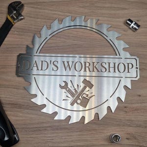 May include: A metal sign shaped like a saw blade, with the words "DAD'S WORKSHOP" cut out. The sign features a tool graphic and is surrounded by tools such as a wrench, pliers, and sockets. The sign is displayed on a wooden surface.