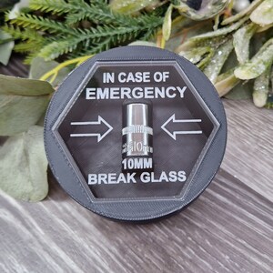 May include: A black hexagonal-shaped container with a clear lid. The lid has a 10mm socket wrench inside and the text "IN CASE OF EMERGENCY 10MM BREAK GLASS" printed on it.
