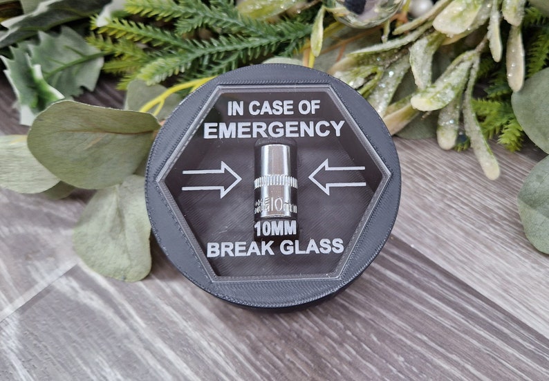 May include: A black hexagonal-shaped container with a clear lid. The lid has a 10mm socket wrench inside and the text "IN CASE OF EMERGENCY 10MM BREAK GLASS" printed on it.