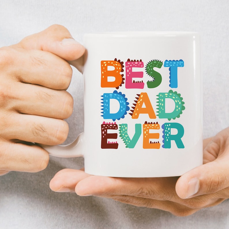 以下が含まれることがあります： 白いセラミック製のマグカップに、カラフルな「BEST DAD EVER」のデザインが施されています。文字は緑色の恐竜のような模様で縁取られています。