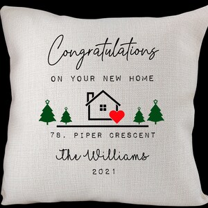 May include: White throw pillow with black text that reads "Congratulations on your new home".  A black silhouette of a house with a red heart is above the text "78. Piper Crescent the Williams 2021".  Two green Christmas trees are on either side of the house.