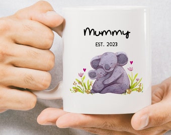 Può includere: Una tazza in ceramica bianca con un'illustrazione in bianco e nero di due koala che si abbracciano. Il testo "Mummy EST. 2023" è stampato sulla tazza.