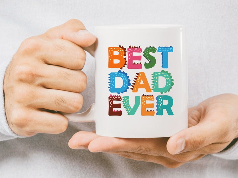 以下が含まれることがあります： 白いセラミック製のマグカップに、カラフルな「Best Dad Ever」のデザインが施されています。文字は恐竜柄で飾られています。