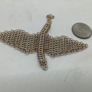 May include: A silver chainmail bird sculpture with outstretched wings. The bird is made of small, interlocking metal rings. A coin is next to the sculpture for size comparison. The bird's wingspan is approximately 10 cm.