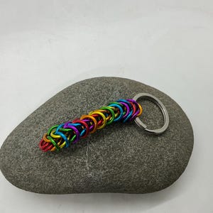 May include: A rainbow-colored chainmail keychain with a silver key ring. The keychain is made of small, brightly colored metal rings linked together in a chain. The keychain is resting on a gray stone.