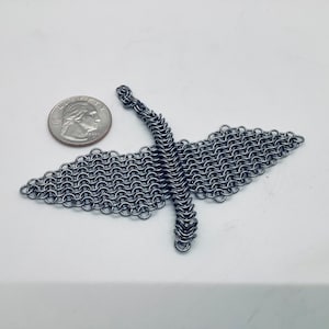 Op de afbeelding: Een handgemaakte kraanvogelsculptuur van maliënkolder, weergegeven op een witte ondergrond, naast een Amerikaanse kwartdollar ter vergelijking. De kraanvogel is gemaakt van kleine, in elkaar grijpende metalen ringen.