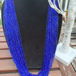 Puede incluir: Un collar de múltiples hilos compuesto por pequeñas y vibrantes cuentas azules. El collar está colocado sobre un maniquí negro, mostrando su diseño en cascada. Las cuentas crean una apariencia rica y texturizada, perfecta para añadir un toque de color.