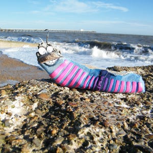 May include: A colourful, handmade sock creature with a smiling face and googly eyes, resting on a rocky surface by the sea. The sock is blue with pink and purple stripes. The background shows the sea and a pier.