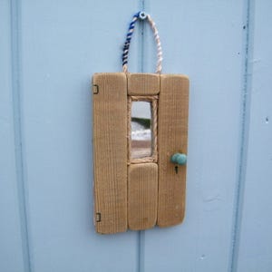 Puede incluir: Un adorno de pared rectangular con forma de puerta en miniatura, construido con tablones de madera verticales de color marrón claro. Presenta un pequeño espejo rectangular central, un pomo redondo azul claro y una cuerda trenzada azul y blanca.