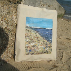 Może przedstawiać: Biała torba z pł&oacute;tna z kolorowym obrazem przedstawiającym scenę plażową. Obraz przedstawia zatłoczoną plażę z ludźmi pływającymi, opalającymi się i bawiącymi się w wodzie. Plaża jest ograniczona przez molo i rząd budynk&oacute;w w oddali. Niebo jest niebieskie, a woda turkusowa.