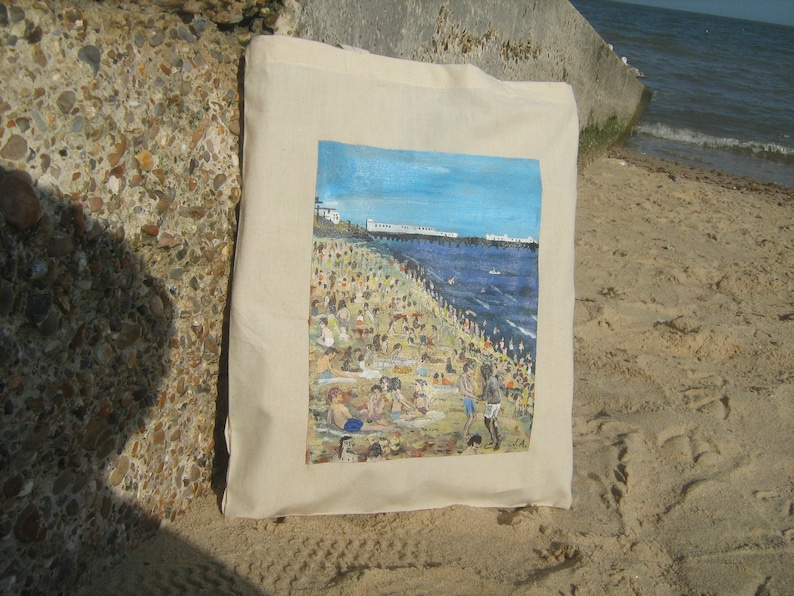 Może przedstawiać: Biała torba z pł&oacute;tna z kolorowym obrazem przedstawiającym scenę plażową. Obraz przedstawia zatłoczoną plażę z ludźmi pływającymi, opalającymi się i bawiącymi się w wodzie. Plaża jest ograniczona przez molo i rząd budynk&oacute;w w oddali. Niebo jest niebieskie, a woda turkusowa.