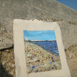 Może przedstawiać: Biała torba z pł&oacute;tna z kolorowym obrazem przedstawiającym scenę plażową. Obraz przedstawia pomost, błękitny ocean i zatłoczoną plażę z ludźmi opalającymi się i kąpiącymi.