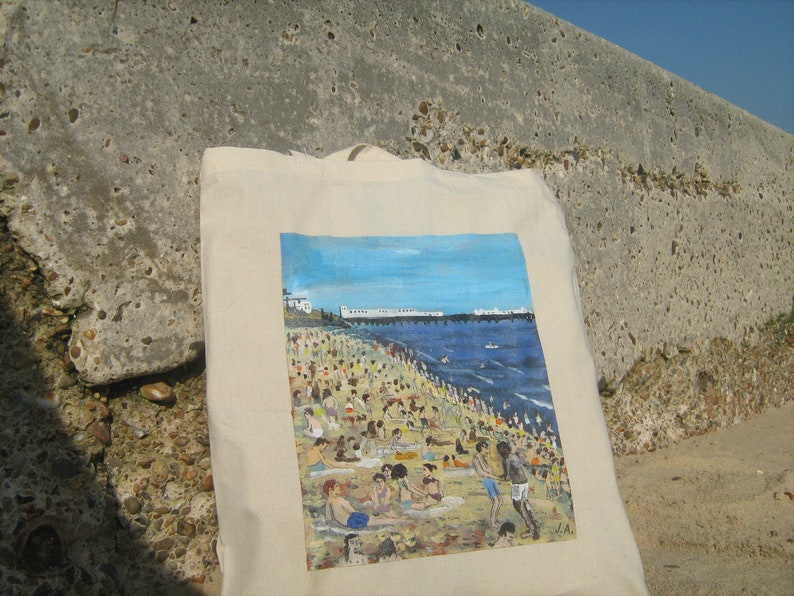 Może przedstawiać: Biała torba z pł&oacute;tna z kolorowym obrazem przedstawiającym scenę plażową. Obraz przedstawia pomost, błękitny ocean i zatłoczoną plażę z ludźmi opalającymi się i kąpiącymi.