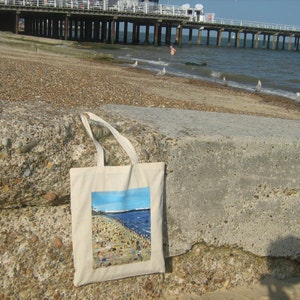 Może przedstawiać: Biała torba z pł&oacute;tna z kolorową ilustracją sceny plażowej. Ilustracja przedstawia ludzi na plaży z molo w tle. Torba ma dwa uchwyty.