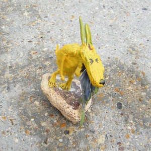 May include: A whimsical, yellow sculpture of a dog-like creature with long ears, holding a dark object. The sculpture is perched on a light-colored, textured rock. The background is a speckled, gray concrete surface.