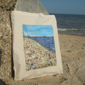 Może przedstawiać: Biała torba z pł&oacute;tna z kolorowym obrazem przedstawiającym scenę plażową. Obraz przedstawia pomost, błękitny ocean i wiele os&oacute;b na plaży.