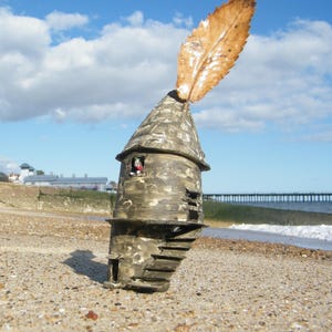 Könnte beinhalten: Ein Miniatur-Turm aus verwittertem Holz in Grau- und Brauntönen mit einem Blatt auf der Spitze. Der Turm hat ein kleines Fenster und eine Wendeltreppe. Der Turm steht auf einem Sandstrand mit einem Steg im Hintergrund.