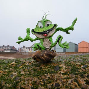 Peut inclure: Une sculpture de grenouille verte fantaisiste, les bras tendus, perchée sur un rocher recouvert de mousse. La grenouille a une expression souriante avec une langue rouge. En arrière-plan, des cabanes de plage colorées et un ciel nuageux.